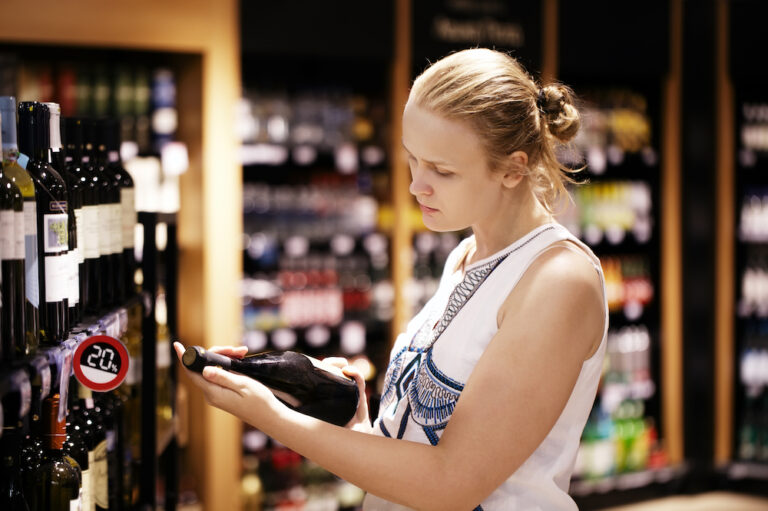 Woman Reading Back of Wine Bottle in Liquor Store, thinking about a wine store pos system